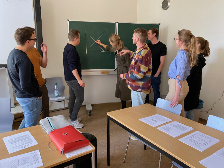 Auf dem Foto stehen drei Referendarinnen und fünf Referendare seitlich und mit dem Rücken zur Kamera um eine grüne Tafel. Die Referendarin im Zentrum zeichnet in ein Koordinatensystem  mit Kreide zwei Geraden ein, die für Angebot und Nachfrage stehen. Der Kurs diskutiert die Implikationen des Schaubildes lebhaft.