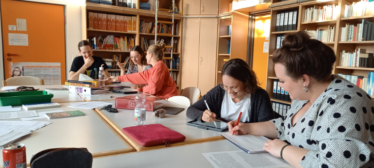 Das Bild zeigt vier Referendarinnen und einen Referendar die in einer Bibliothek an einem großen Tisch sitzen und mit Texten arbeiten. Teilweise arbeiten sie dabei mit analogen, teilweise mit digitalen Medien. Auf dem Tisch liegen verschiedene Unterrichtsmaterialien und Bücher aus dem Bereich Ethik/Philosophie.