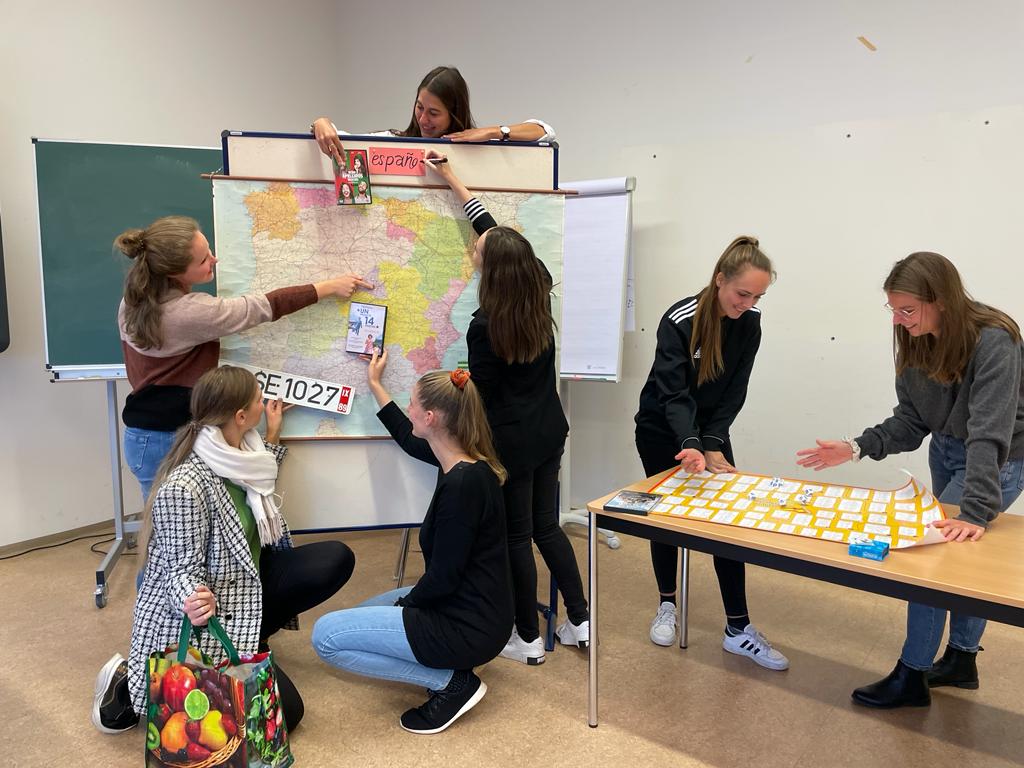 Die Referendare befinden sich in einem Raum des Seminars. Insgesamt sind es sieben Referendarinnen. Im Hintergrund sieht man eine Landkarte von Spanien an einer Pinnwand und die Referendarinnen sind um diese positioniert. Eine Referendarin steht hinter der Pinnwannd und blickt von hinten auf die Landkarte. In ihrer Hand hält sie eine DVD des Films „Ocho apellidos vascos“, ein spanischer Film. Dabei hält sie die DVD auf das Baskenland gerichtet.  Im vorderen Bereich befinden sich vier Referendarinnen, davon zwei auf der linken Seite und zwei auf der rechten Seite. Von denen auf der linken Seite hält eine Referendarin eine Obsttasche in der Hand und ein Kennzeichen von Sevilla, zeigt auf die Stadt in Andalusien und kniet auf dem Boden, die andere zeigt mit ihrer rechten Hand auf Madrid und steht dahinter.  Auf der rechten Seite hält eine Referendarin die DVD „Un Franco, 14 pesetas“ in der Hand und blickt in dieselbe Richtung. Sie kniet ebenfalls. Die vierte Referendarin steht und schreibt auf eine rote Karte español.  Zwei weitere Referendarinnen stehen rechts von der Landkarte an einem Tisch, blicken auf eine mindmemo, auf der sich spanischer Wortschatz befindet und würfeln storycubes, mit denen man im Unterricht Geschichten erzählen kann. Insgesamt schauen alle Referendarinnen sehr freundlich.