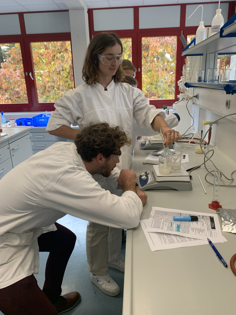 Das Bild zeigt vier Chemie-Referendarinnen und Referendare im Labor. Sie tragen weiße Kittel und Schutzbrillen. Eine Referendarin erwärmt eine klare und eine trübe Flüssigkeit auf einem elektrischen Magnetrührer mit Heizplatte und misst deren Temperaturen mithilfe von Thermometern. Ein Referendar beugt sich nach unten und liest die Temperatur ab. Auf dem Labortisch liegen weitere Arbeitsutensilien, Blätter und Stifte.