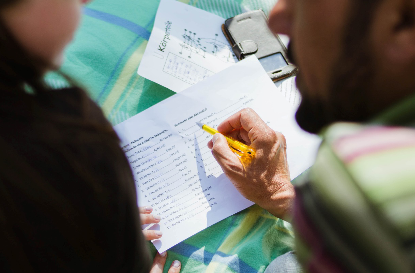 Das Bild zeigt einen Lehrer und eine Schülerin von oben. Auf dem Tisch liegen Arbeitsblätter. Der Lehrer erklärt der Schülerin etwas.