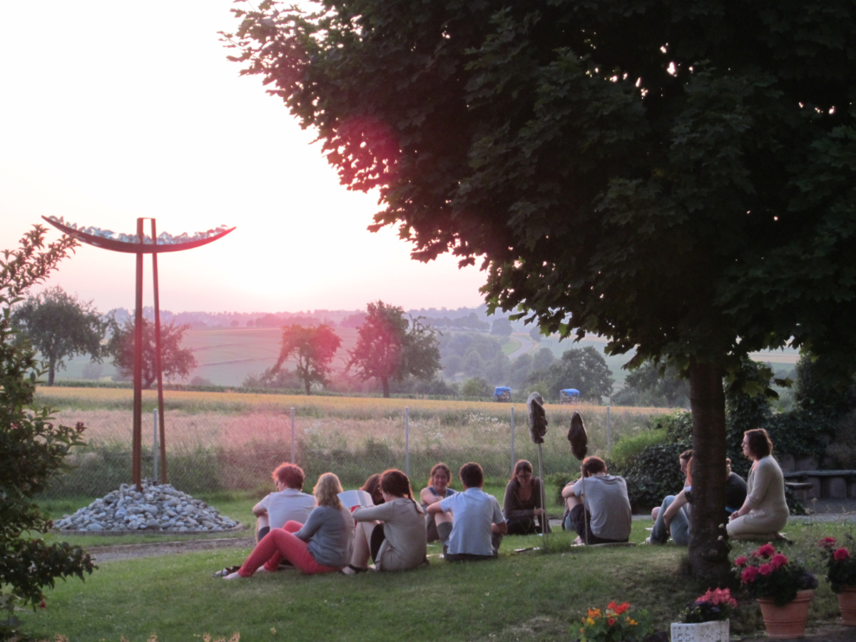 Das Bild zeigt eine Gruppe von Referendarinnen und Referendaren, die in einem Garten zwischen einem Baum und einem großen, modernen, aus Stahl und Steinen gefertigten Kreuz sitzen. Manche lesen, andere blicken in die Ferne, eine Referendarin lächelt. Im Hintergrund sind Wiesen, Obstbäume und Berge zu erkennen. Das Bild ist durch das Abendrot in einen rötlich-goldenen Schimmer getaucht.