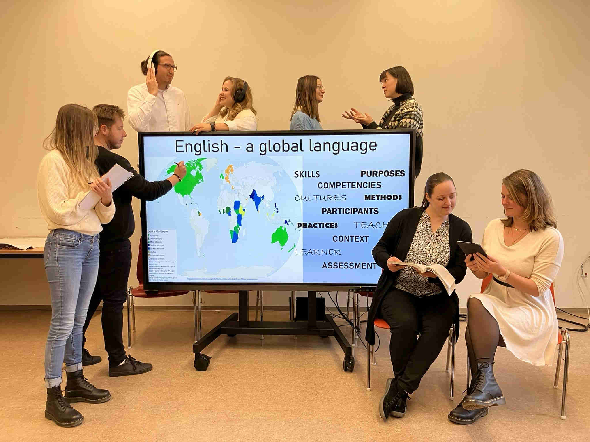 Präsentation zum Thema „Englisch als globale Sprache“ Abgebildet auf dem Bild ist eine Gruppe von sechs Lehrkräften in Ausbildung (LkiA), die an einer Präsentation zum Thema „Englisch als globale Sprache“ gemeinsam arbeiten. Auf einem Bildschirm sind dabei farbige Weltkarten und Begriffe wie 'Skills', 'Competencies' und 'Assessment' zu sehen. Zwei weibliche LkiA sitzen dabei und lesen sich ein, während andere LkiA stehen und diskutieren.