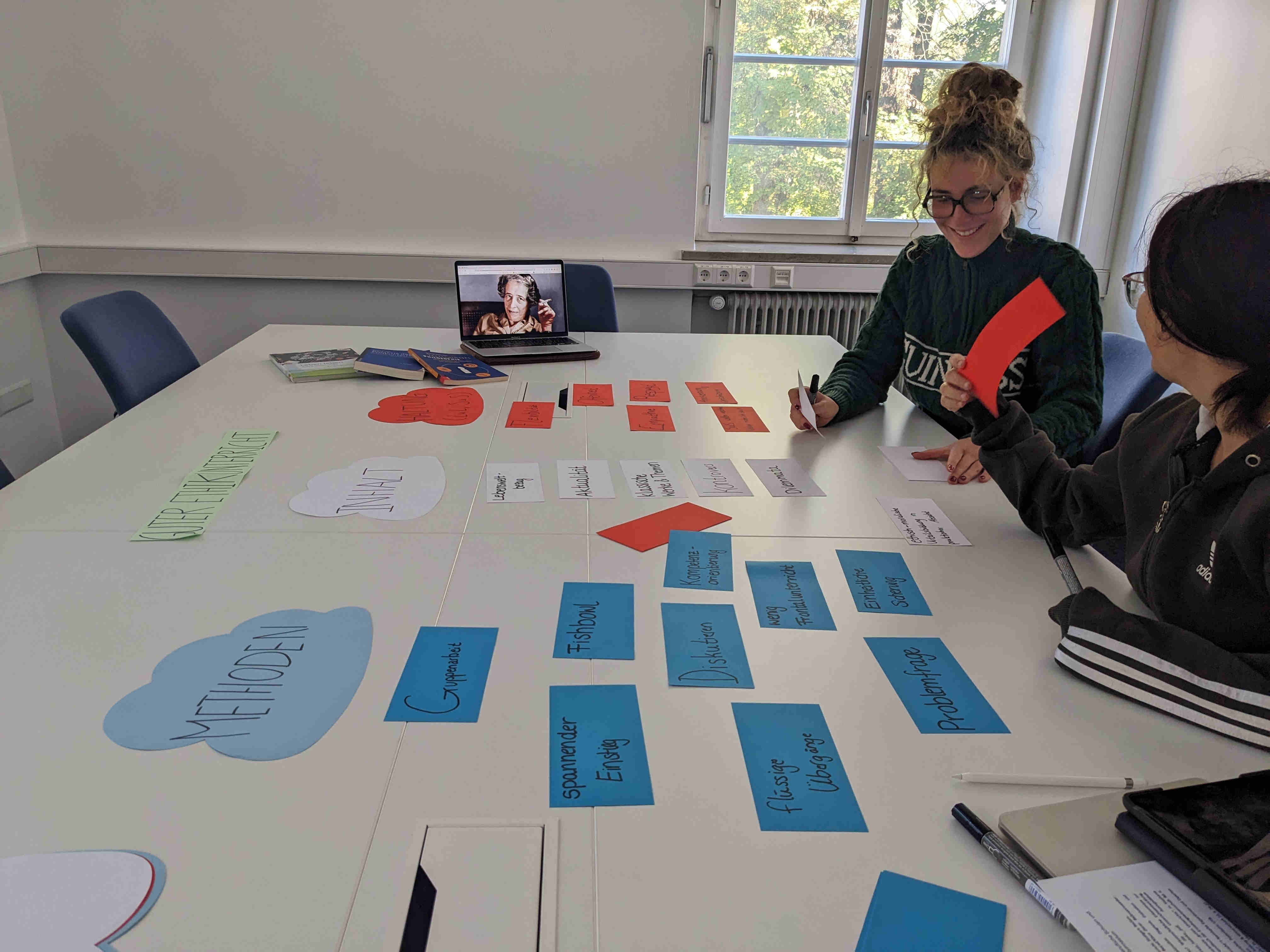 Zu sehen ist ein heller Seminarraum mit einem großen weißen Tisch, auf dem farbige Karteikarten und Papierwolken ausgebreitet sind. Auf den Karten stehen verschiedene Begriffe wie „Methoden“, „Inhalt“ und „Gruppenarbeit“. Zwei Personen sitzen am Tisch und arbeiten gemeinsam, sie schreiben auf Karten und lächeln. Im Hintergrund ist ein geöffnetes Fenster, durch das Tageslicht hereinfällt. Auf dem Tisch steht außerdem ein Laptop, auf dessen Bildschirm eine Person zu sehen ist. Die Szene wirkt konzentriert, aber freundlich und kooperativ – typisch für eine Gruppenarbeit oder Unterrichtsvorbereitung.