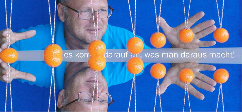 Auf der bildlichen Darstellung ist ein freundlich und zugleich nachdenklich blickender Mann mit Brille zu sehen. Dieser befindet sich positioniert vor einer Konstruktion mit orangefarbenen Bällen, die an Schnüren hängen und vor einem blauen Hintergrund zu sehen sind. Zwischen den Bällen befindet sich ein Text: 'Es kommt darauf an, was man daraus macht!'. Der Herr auf dem Bild ist der Fachleiter für die Fachdidaktik Physik am Seminar Weinarten, Herr Studiendirektor Wolfgang Kreitmair.