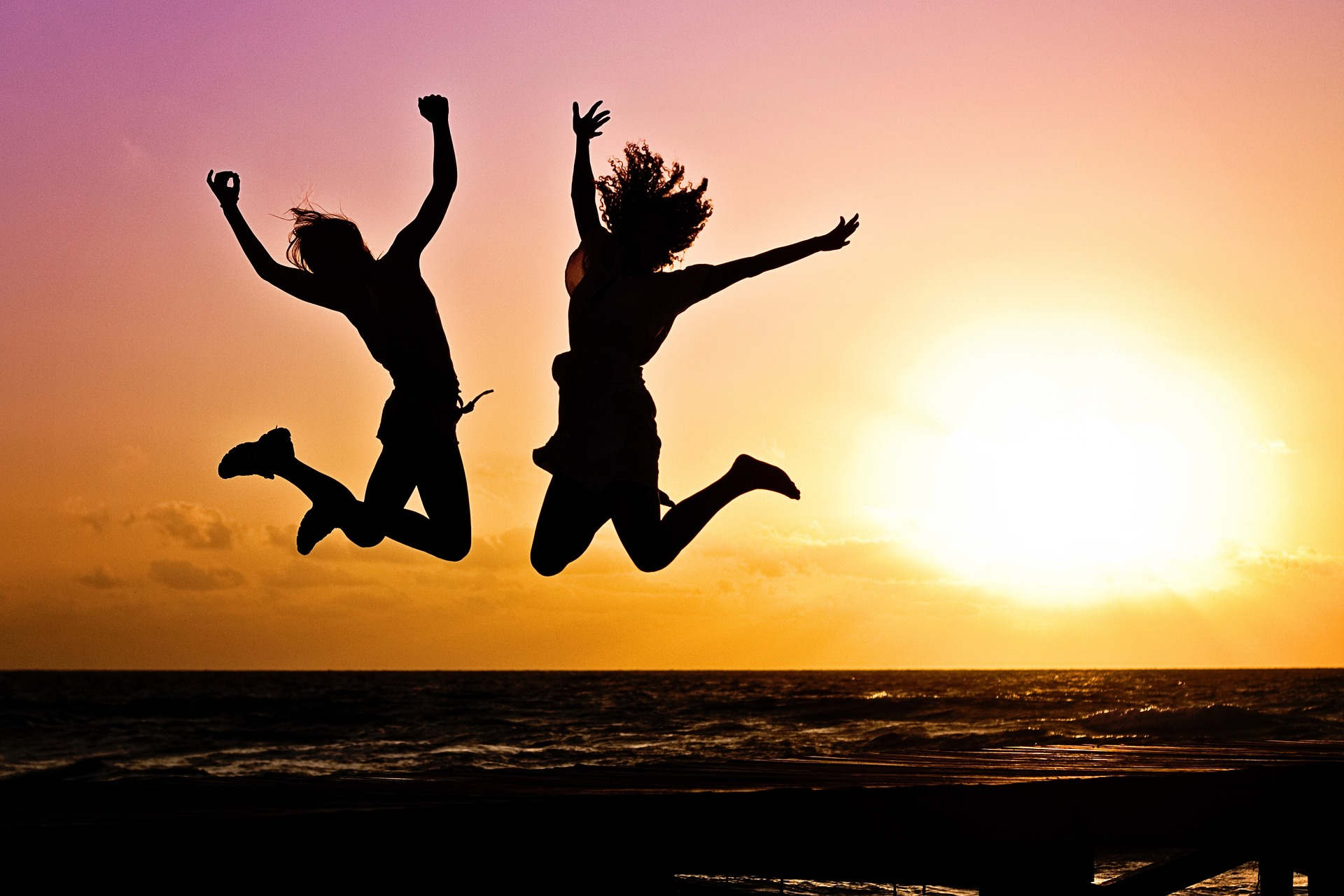 Zwei Personen, die vor einem Sonnenuntergang in die Luft springen Auf der bildlichen Darstellung befinden sich die Silhouetten von zwei Personen, die vor einem Sonnenuntergang am Strand hoch in die Luft springen. Im Hintergrund abgebildet ist ein Meer in Abendstimmung. Das Bild symbolisiert positive Emotionen wie Zuversicht, Freude, Freiheit und Hoffnung.