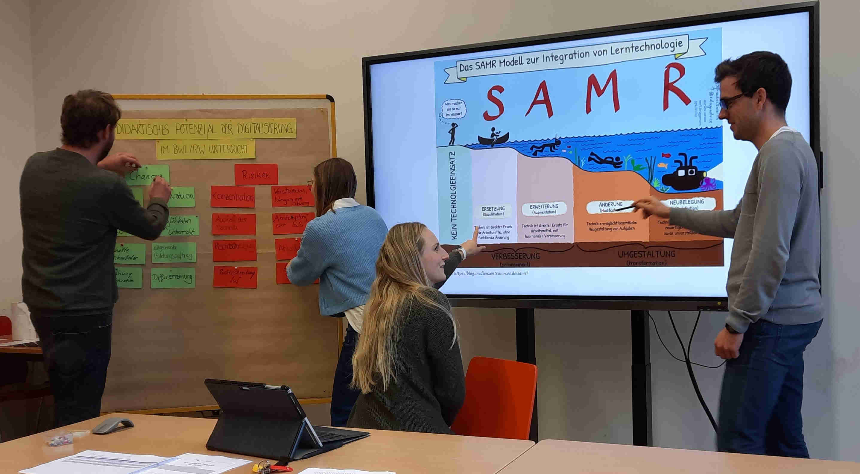 Auf der bildlichen Darstellung sind eine digitale Tafel mit dem SAMR-Model zur Integration von Lerntechnologien und zwei Lehrkräften in Ausbildung zu sehen, die darauf zeigen und sich darüber unterhalten. Daneben steht eine Metaplanwand mit dem Titel „didaktisches Potenzial der Digitalisierung im BWL/RW Unterricht“ und zwei Lehrkräfte in Ausbildung pinnen Karten an die Metaplanwand.
