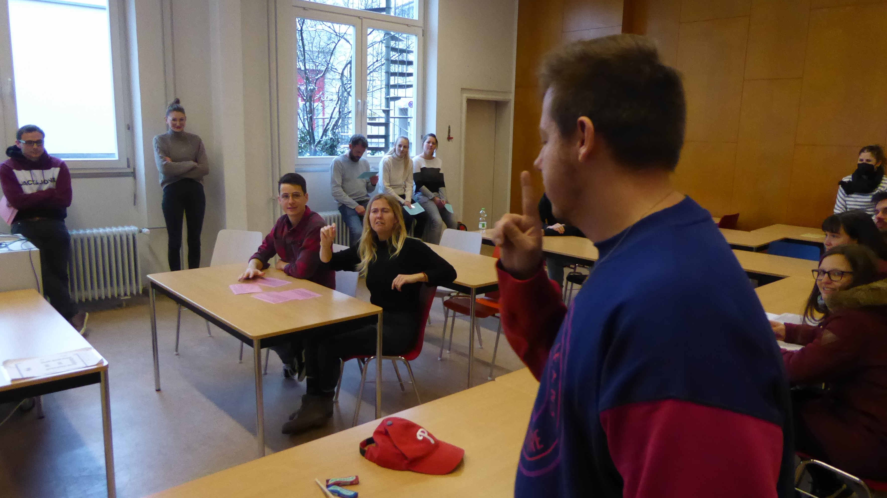 Teambuilding-Maßnahme - Tower of Power Auf der bildlichen Darstellung ist eine Gruppe von Lehrkräften in Ausbildung (LkiA) in einem Raum des Seminares Weingarten abgebildet. Ein Mann spricht mit erhobener rechter Hand und zeigt dabei seinen Zeigefinger, während zwei Personen am Tisch aufmerksam zuhören. Im Hintergrund sind weitere LkiA sichtbar. Dargestellt ist eine Team-Building-Maßnahme mit dem Tower of Power – Geschicklichkeitsspiel, bei dem eine Gruppe versuchen muss, einen Turm aus unterschiedlichen Holzklötzen mittels eines Hakens zu bauen, der über lange Schnüre der Teilnehmenden ringförmig von außen zu bedienen ist.