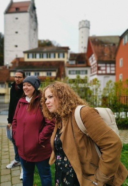 Auf dem Bild zu sehen ist eine Gruppe von drei Lehrkräften in Ausbildung vor historischen Gebäuden der Stadt Ravensburg. Eine Dame mit lockigem Haar und braunem Mantel beugt sich nach vorne, während eine andere Dame in rotem Mantel und Mütze lächelt. Im Hintergrund ist ein Herr mit einem schwarzen Mantel und dunklem Haar abgebildet sowie Türme, Fachwerkhäuser bzw. weitere historischen Bauten zu erkennen.