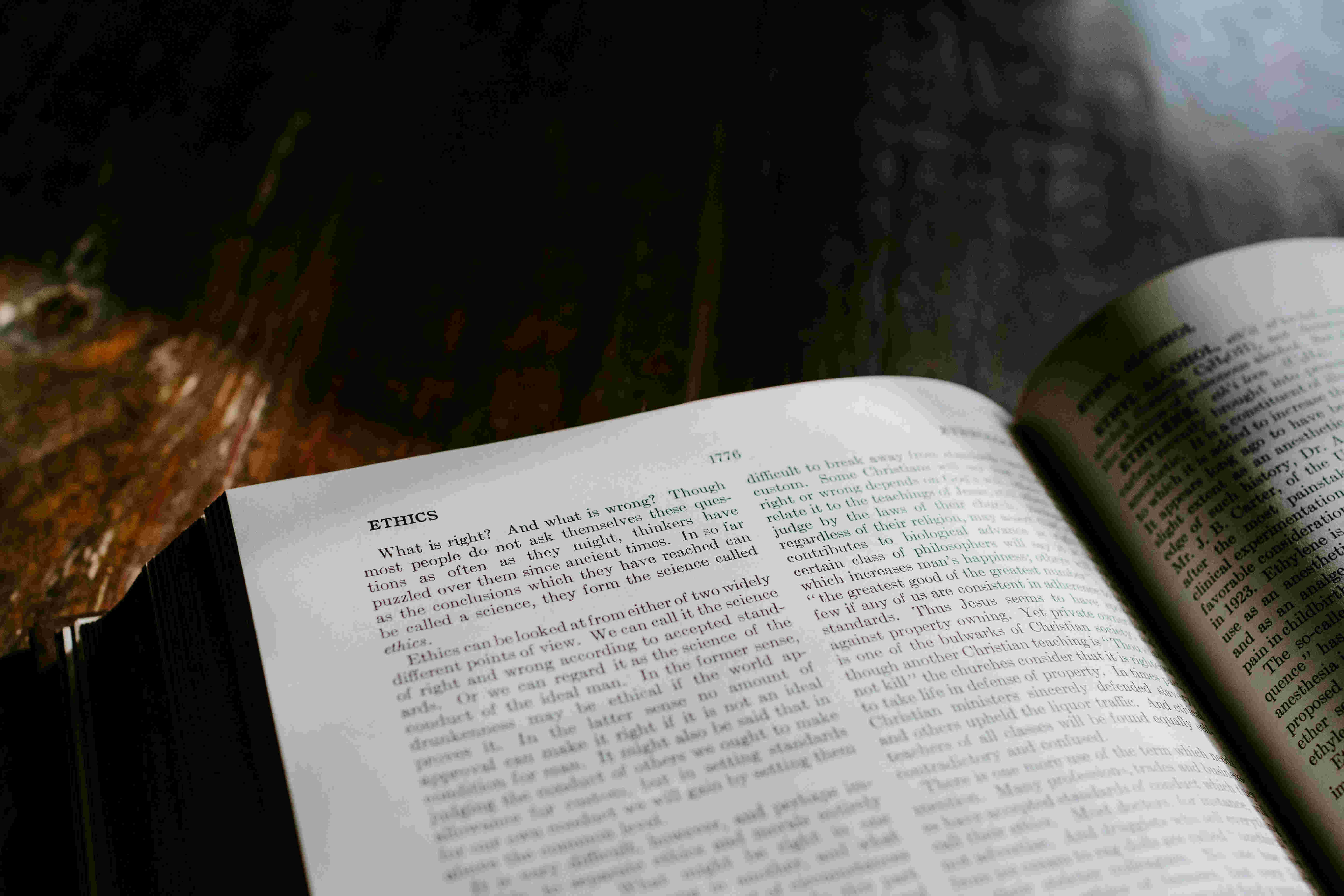 Ethik - eine Definition Auf dem Bild zu sehen ist ein aufgeschlagenes Buch auf einem dunklen Holztisch. Die Seite ist dem Thema „Ethik“ gewidmet und beginnt mit der Frage „Was ist richtig? Und was ist falsch?“. Das Licht fällt schräg von der Seite auf die Buchseite und hebt den gedruckten Text hervor. Der Hintergrund ist unscharf und dunkel, wodurch der Fokus ganz auf dem Buch liegt. Es handelt sich um eine ruhige, nachdenkliche Szene – ideal für konzentriertes Lesen oder wissenschaftliches Arbeiten