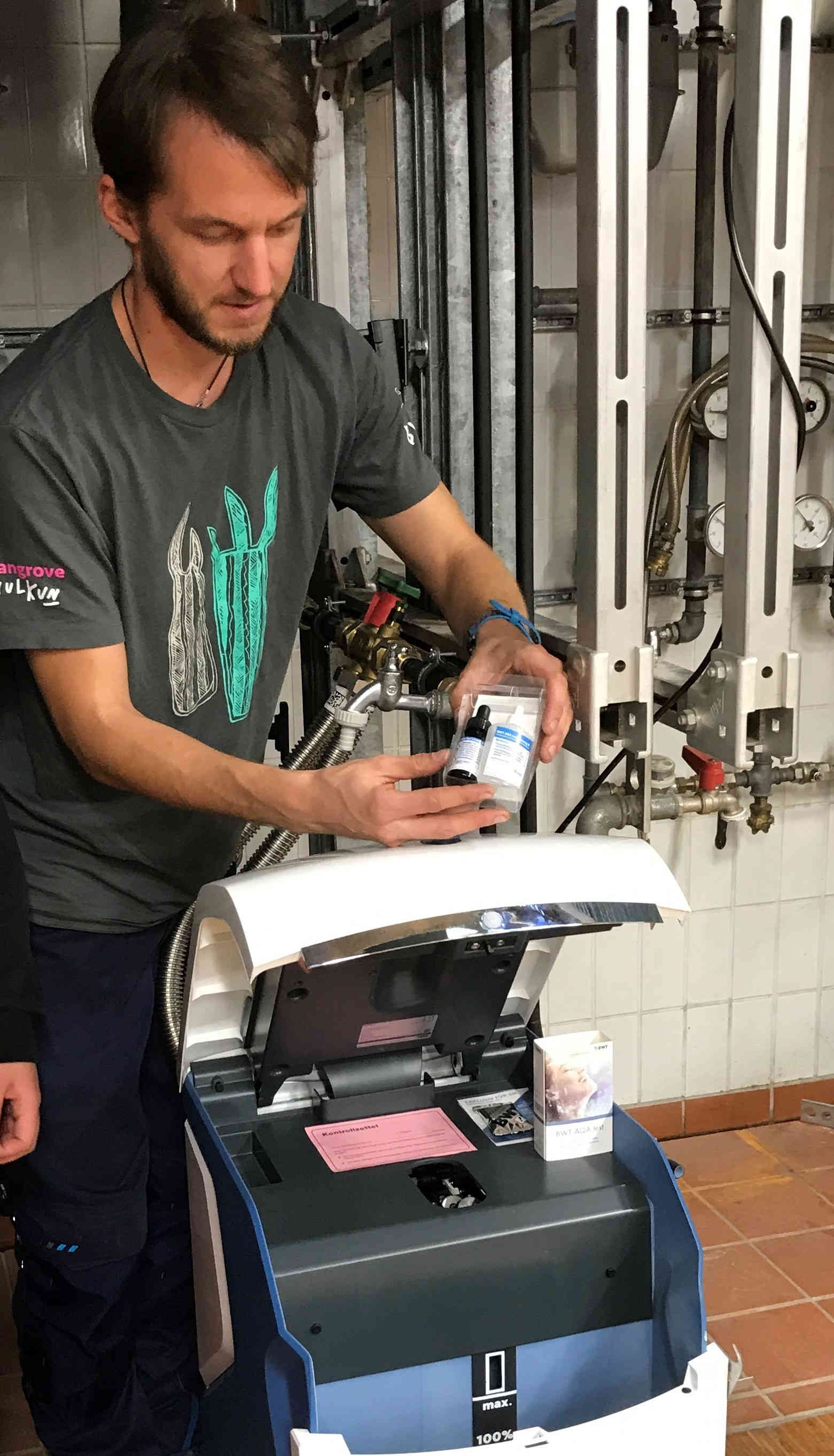 Anlagenmechaniker Auf der Darstellung ist ein Mann in einem grauen T-Shirt, mit braunen Haaren und ebenso braunem Bart abgebildet. Er bedient eine Maschine und hält gleichzeitig ein Kistchen mit mehreren darin enthaltenen Fläschchen in der Hand, welche beispielsweise zur Analyse von Proben dienen könnten. Die abgebildete Szene spielt in einem technischen Raum in dessen Hintergrund mehrere Leitungen und Messelemente sichtbar sind.
